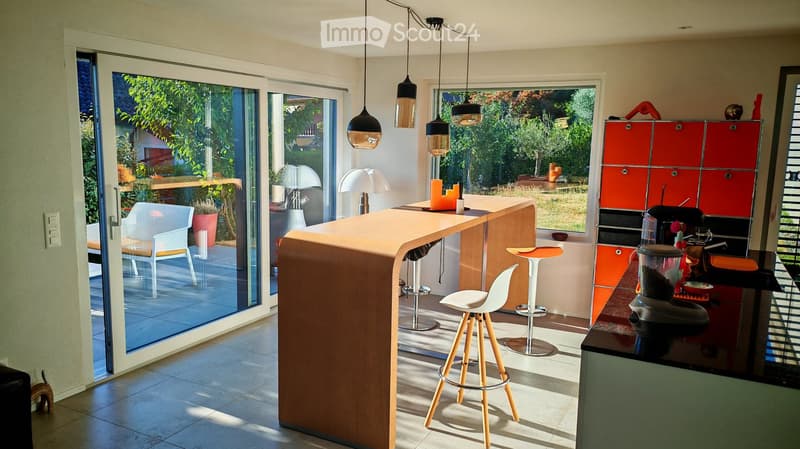 Modern kitchen with a wooden bar counter, white stools, pendant lamps, and red cabinets. Large windows offer a view of the garden.
