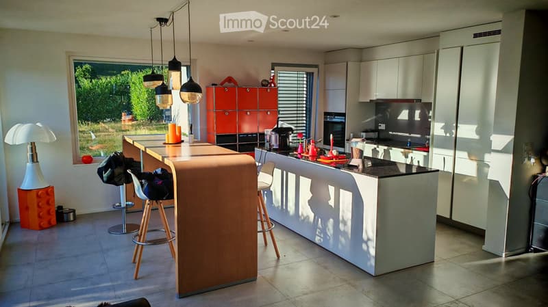 Modern kitchen with white cabinets, a central island, bar stools, and various appliances. Features a large window with a view of greenery outside.