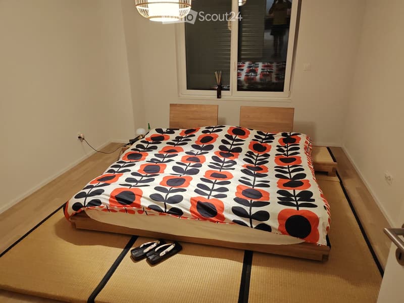 A small bedroom with a wooden bed, colorful bedding, a nightstand with a lamp, and a window with blinds. Shower slippers are placed on the floor.
