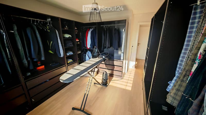 Well-organized walk-in closet with dark wooden wardrobe, shelves, and an ironing board with iron. Clothing is neatly hung and stored.