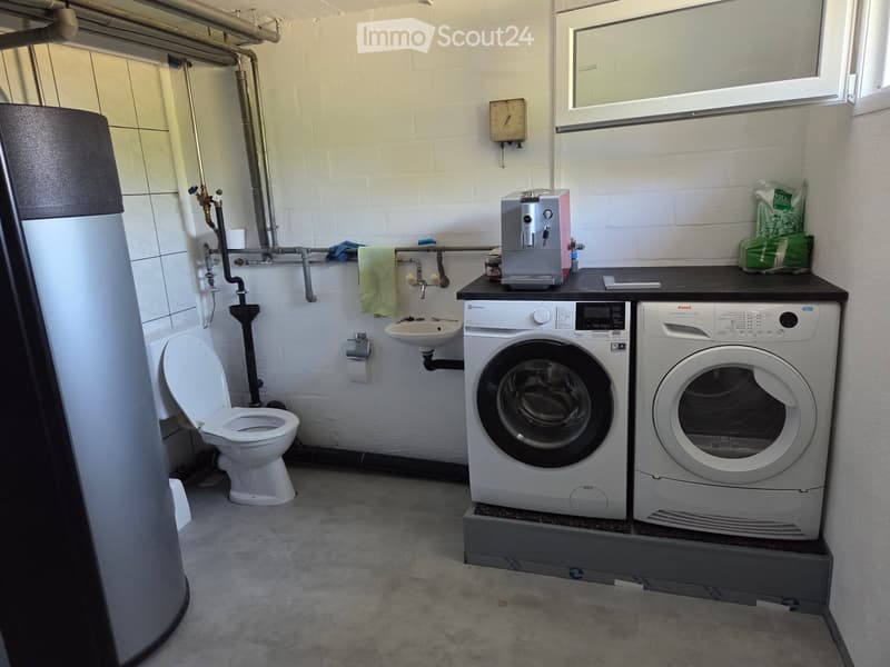 Functional bathroom with a toilet, sink, washing machine, and dishwasher. Includes a towel, coffee machine, and a small window.