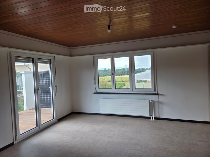 Empty room with white walls, wooden ceiling, large windows, and a sliding glass door leading to a wooden deck.