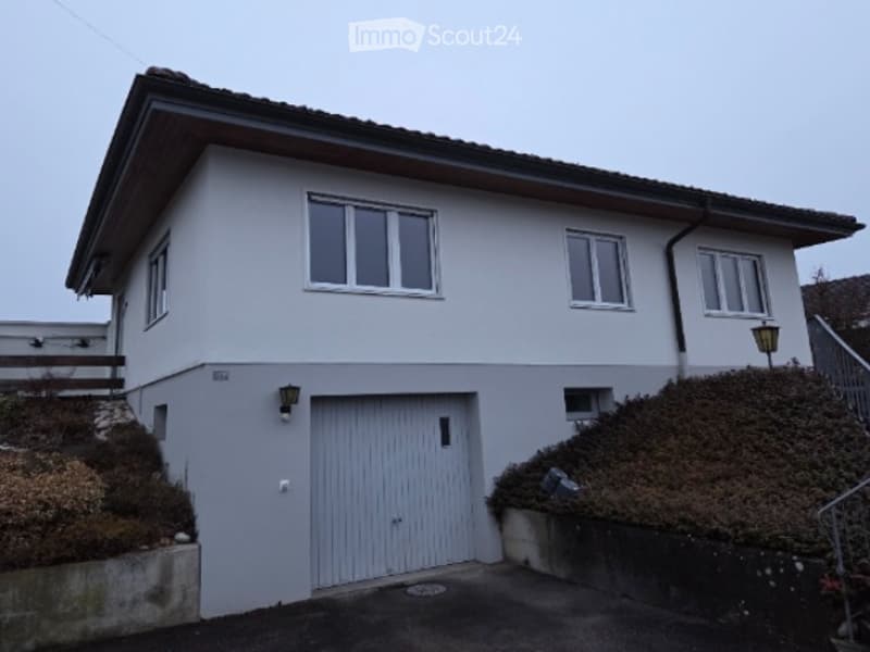 Two-story house with white exterior, grey garage door, multiple windows, and a small garden area.