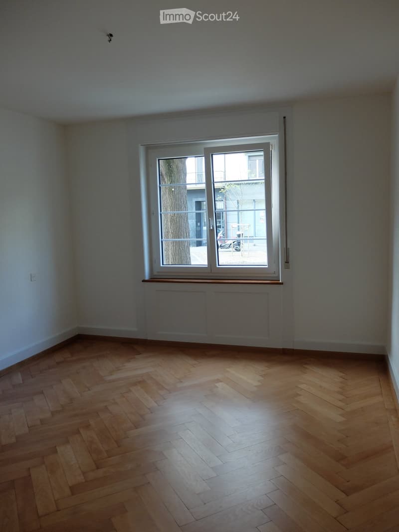 Empty room with wooden herringbone flooring, white walls, and a large window with a view of a tree and balcony area.