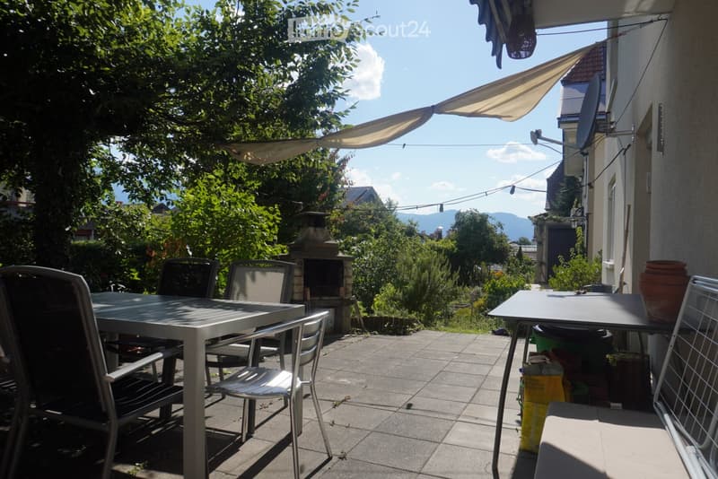 Outdoor patio with a table, chairs, and a barbecue, surrounded by greenery and a clear sky.