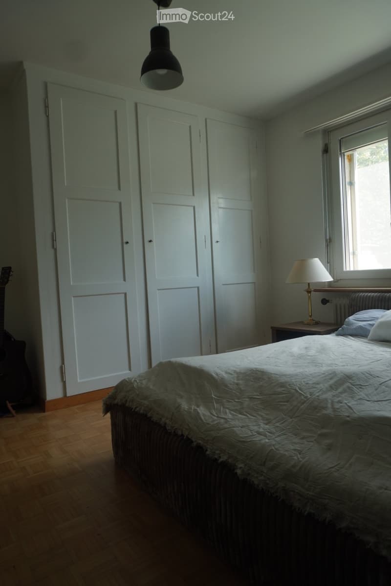 Bedroom with wooden floor, bed with white bedding, guitar leaning against wall, nightstand with lamp, and three white doors.