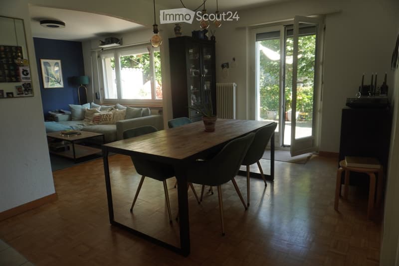 Bright living and dining area with wooden flooring, large windows, and a sliding glass door leading to a balcony. Features a grey sofa, coffee table, dining table with chairs, and a tall cabinet.
