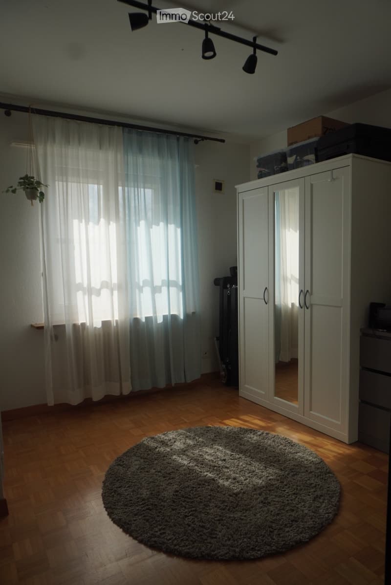 Bright room with wooden flooring, large window with sheer curtains, round shaggy rug, white wardrobe with mirrored doors, and a hanging plant.