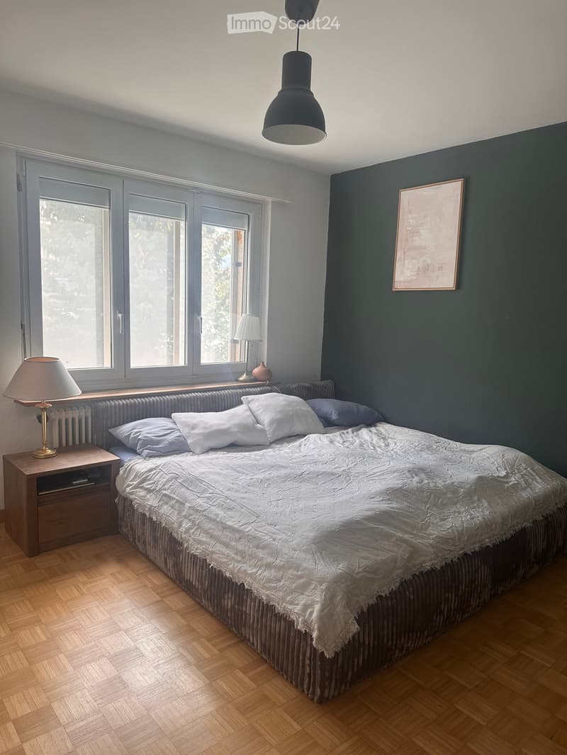 Bedroom with a large bed, nightstand, lamp, and picture frame on the wall. The room features wooden flooring and large windows with blinds.