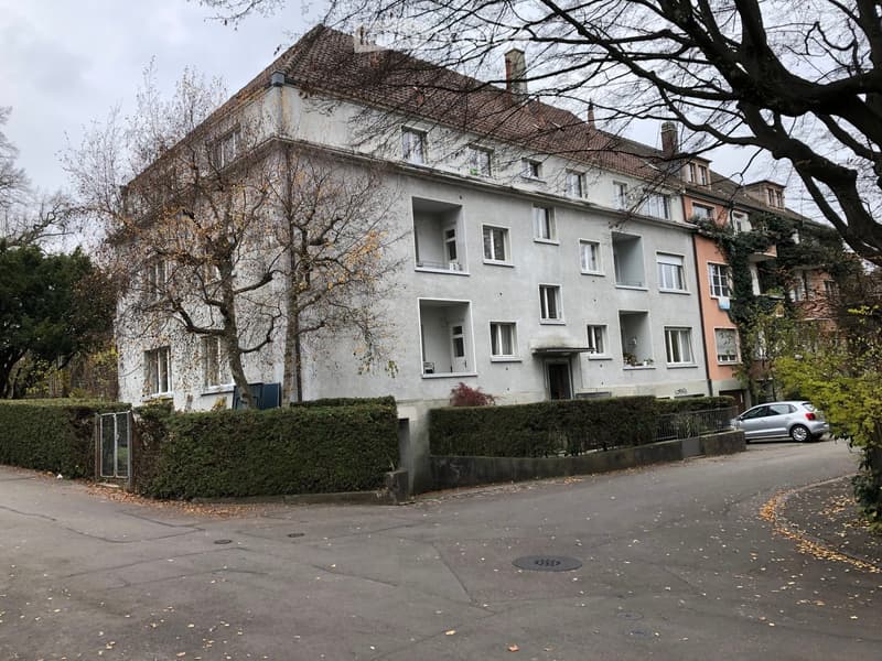 A multi-story residential building with white walls, multiple windows, and balconies, surrounded by trees and a parked car.