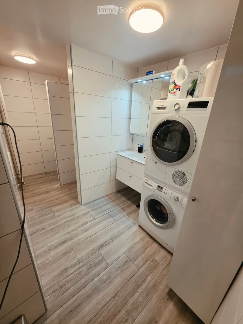 Modern bathroom with wooden flooring, white tiled walls, a washing machine, a sink, and a built-in cupboard.
