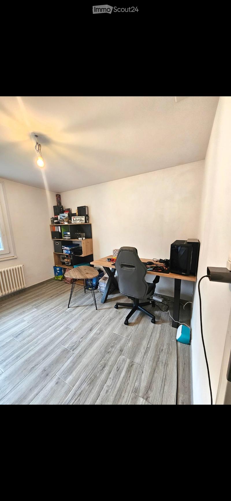 A modern, well-lit room with a wooden floor, a large desk with a chair, a small round stool, a shelf with books and electronic devices, and a window letting in natural light.