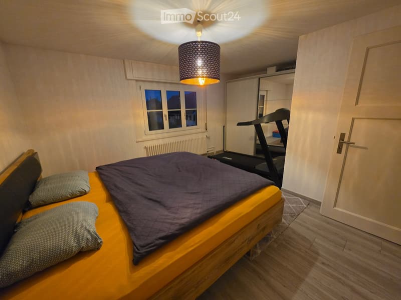 Cozy bedroom with a wooden bed, dark blue duvet, patterned pillows, pendant light, wardrobe, and a treadmill in the corner.