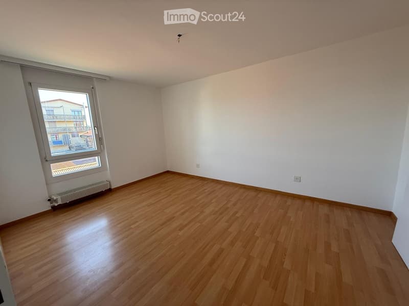 Empty room with wooden flooring, white walls, and a large window with a view of a building and balcony.