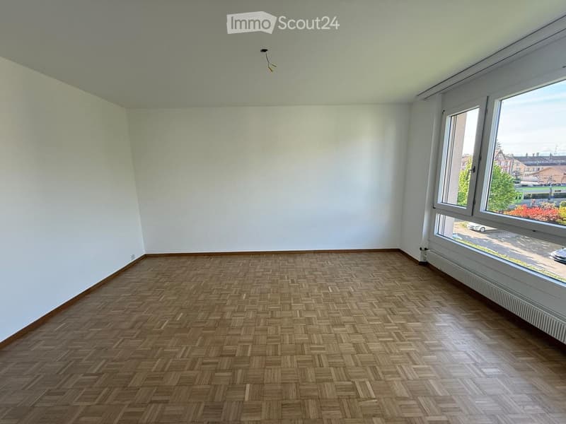 Empty room with wooden herringbone flooring and white walls, featuring large windows with a view of the street.