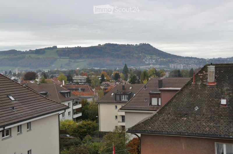 Residential neighborhood with multiple houses featuring red-tiled roofs, surrounded by trees and a distant hilly landscape under an overcast sky.