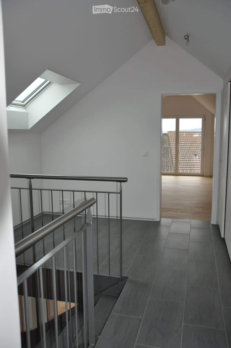 Modern hallway with grey tiled floor, metal railing staircase, skylight window, and an open doorway leading to another room with wooden flooring.