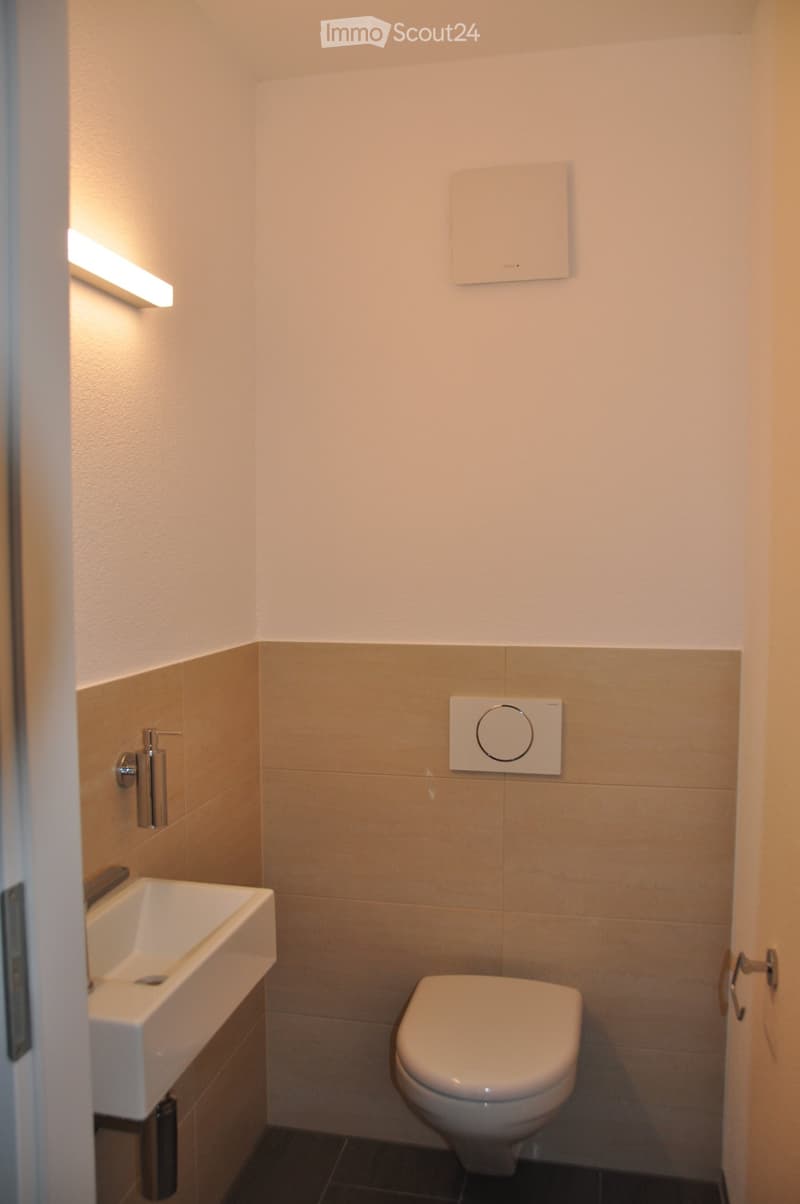Modern bathroom with a white sink, toilet, and a soap dispenser mounted on the wall. The walls are tiled in beige and white, with a sleek, minimalist design.