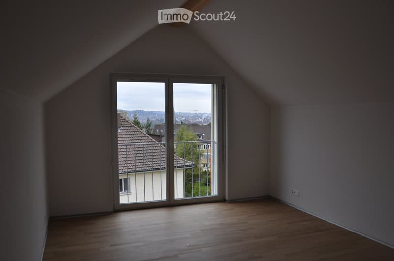 Empty attic room with wooden flooring, large sliding glass door leading to a balcony, and exposed wooden beams on the ceiling.