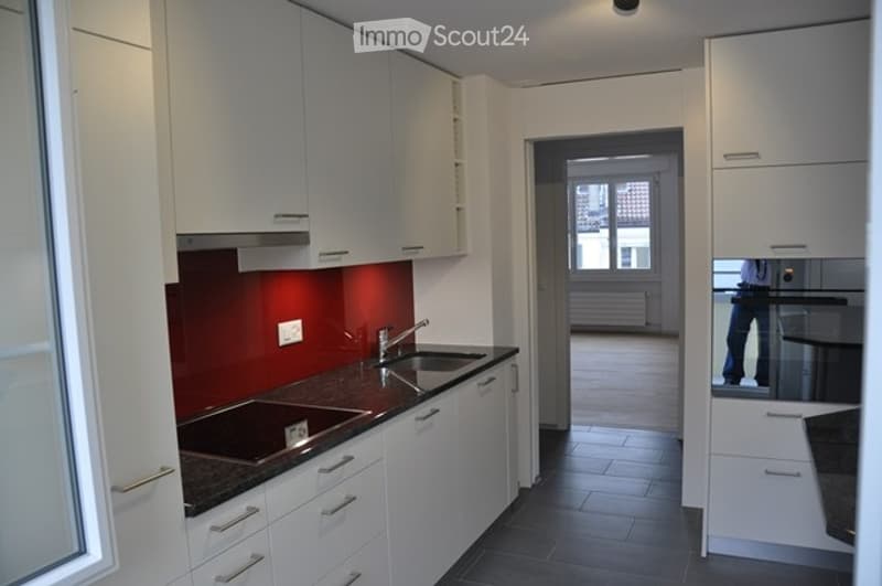 Modern kitchen with white cabinets, dark countertops, and a red accent wall. Features a sink with a modern faucet and a visible hallway leading to another room.