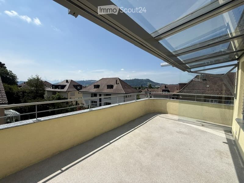 Balcony with a modern glass roof, overlooking a residential area with houses and mountains in the background.
