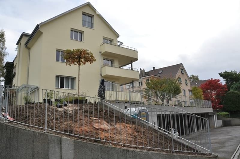 A multi-story residential building with a raised ground floor, balcony, garage, and surrounding greenery.