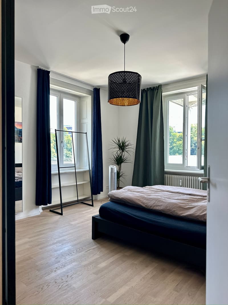 Modern bedroom with wooden flooring, large windows, dark curtains, a bed with beige bedding, a hanging lamp, a mirror, a plant, and a radiator.