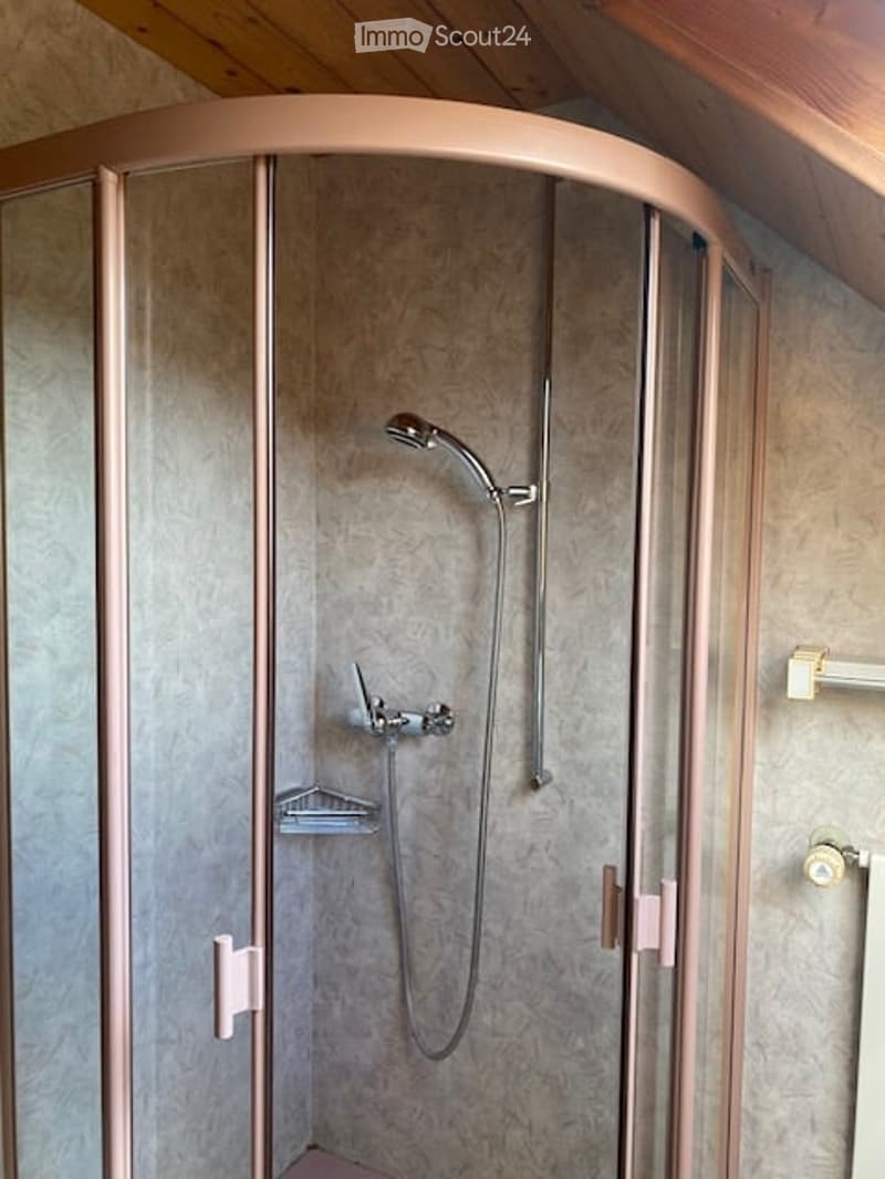 Modern shower stall with a curved glass enclosure, a handheld showerhead, and a built-in seat. The walls are tiled in a light, textured pattern, and the enclosure features rose-gold handles and fixtures.