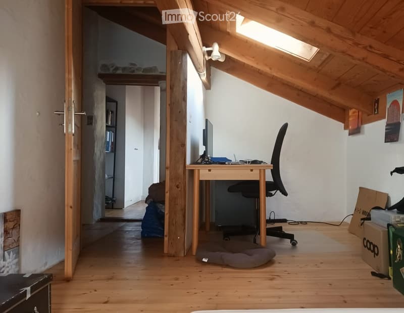 A small, furnished room with wooden flooring, a desk with a screen, a chair, a pet bed, and a lamp on the wall. The room has slanted wooden ceilings and an open doorway leading to another area.
