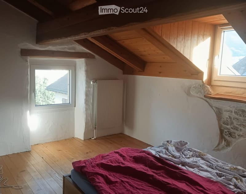 Cozy attic bedroom with wooden floors, exposed wooden beams, white walls, and two windows offering natural light. Features a bed with a red blanket and a radiator on the wall.