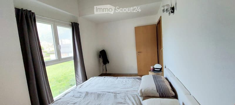 Bedroom. A small, well-lit bedroom with a single bed, pillows, a nightstand with a hat, and a curtained window overlooking a green area.