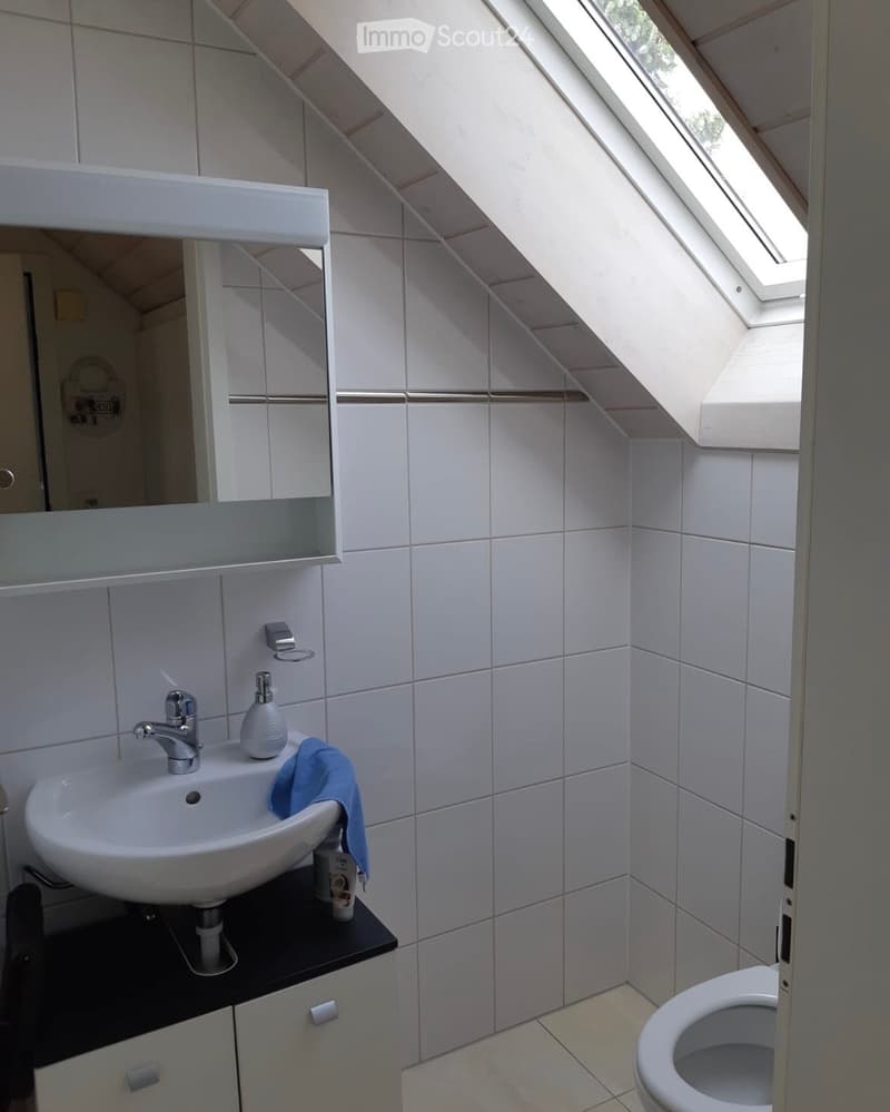 Modern bathroom with white tiles, a sink with a modern faucet, a mirror cabinet, and a toilet. The room features a skylight window and a small black countertop with a cup and a blue towel.