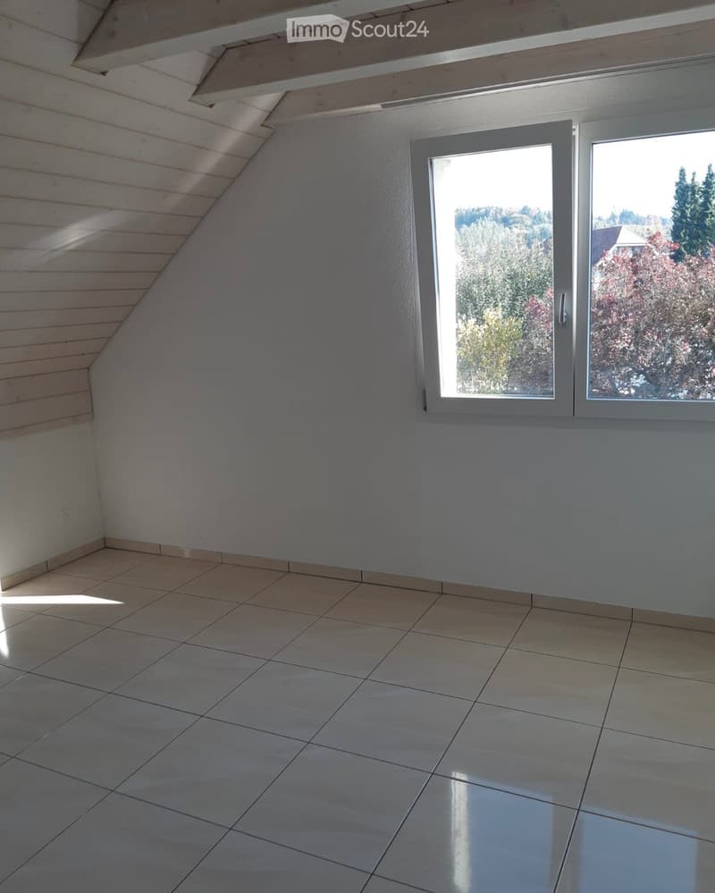 Empty room with tiled floor, white walls, and large windows offering a view of trees and a house outside.