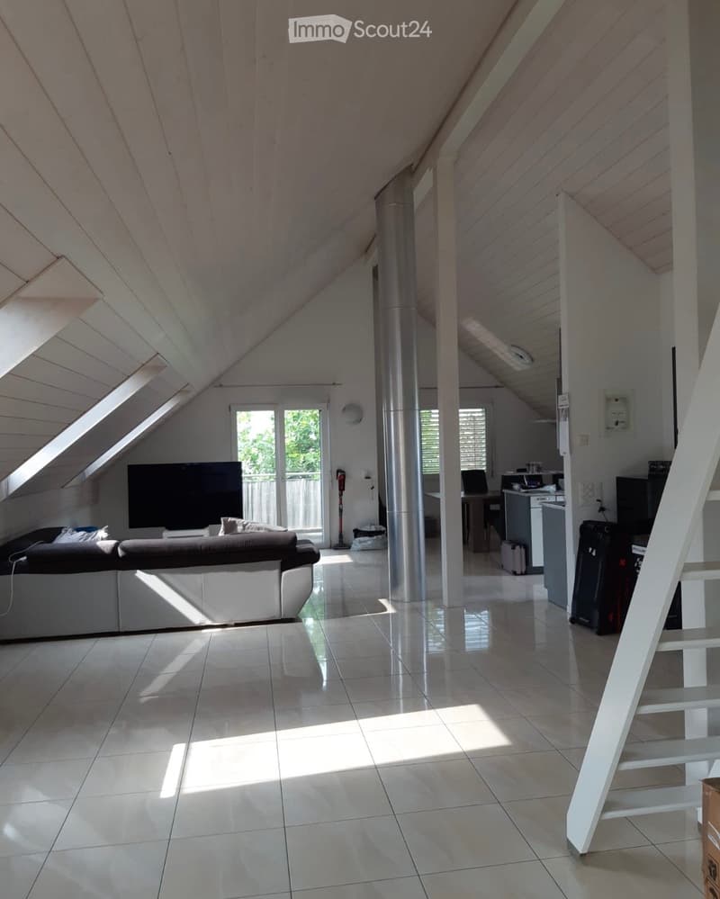 Bright, modern living space with a slanted wooden ceiling, large windows, and a sofa facing a television. The room includes a small kitchen area with a table and chairs, a laptop on a desk, and a suitcase near the entrance.
