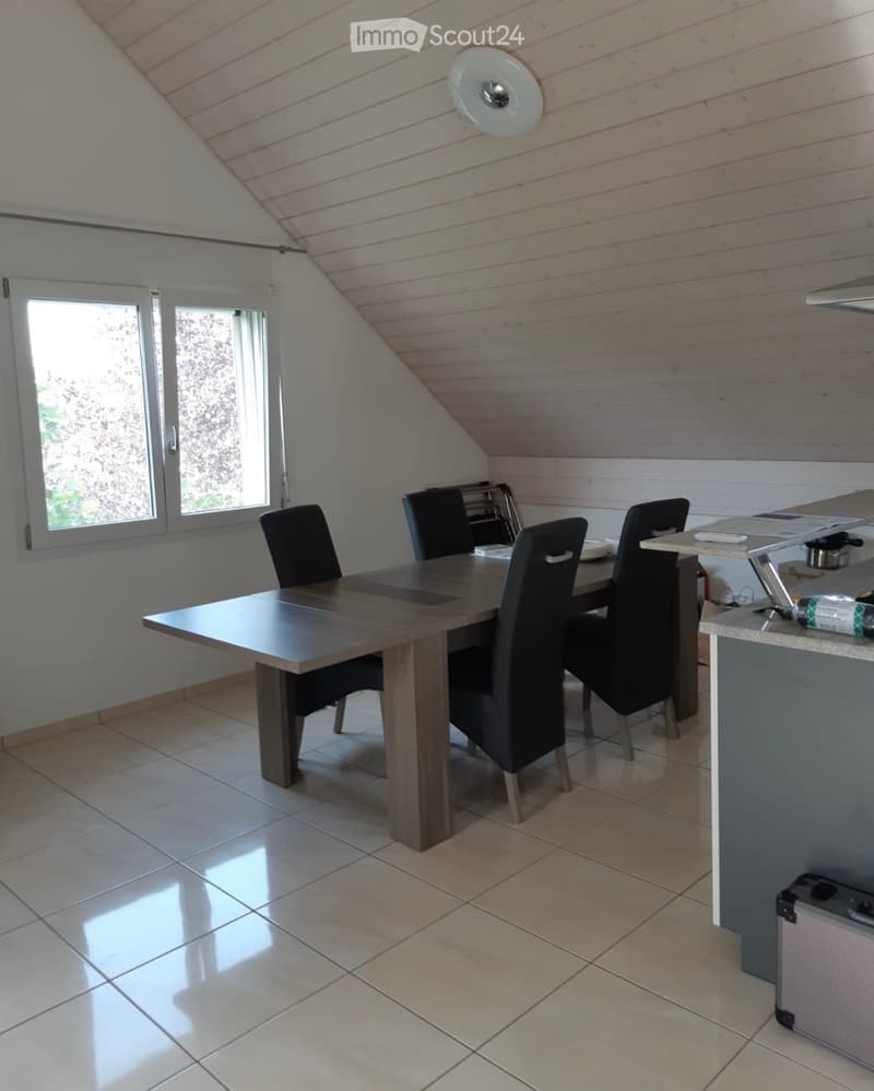 Bright dining area with a wooden dining table, four black chairs, and a kitchen counter with a small appliance. The room has a slanted wooden ceiling and large windows letting in natural light.
