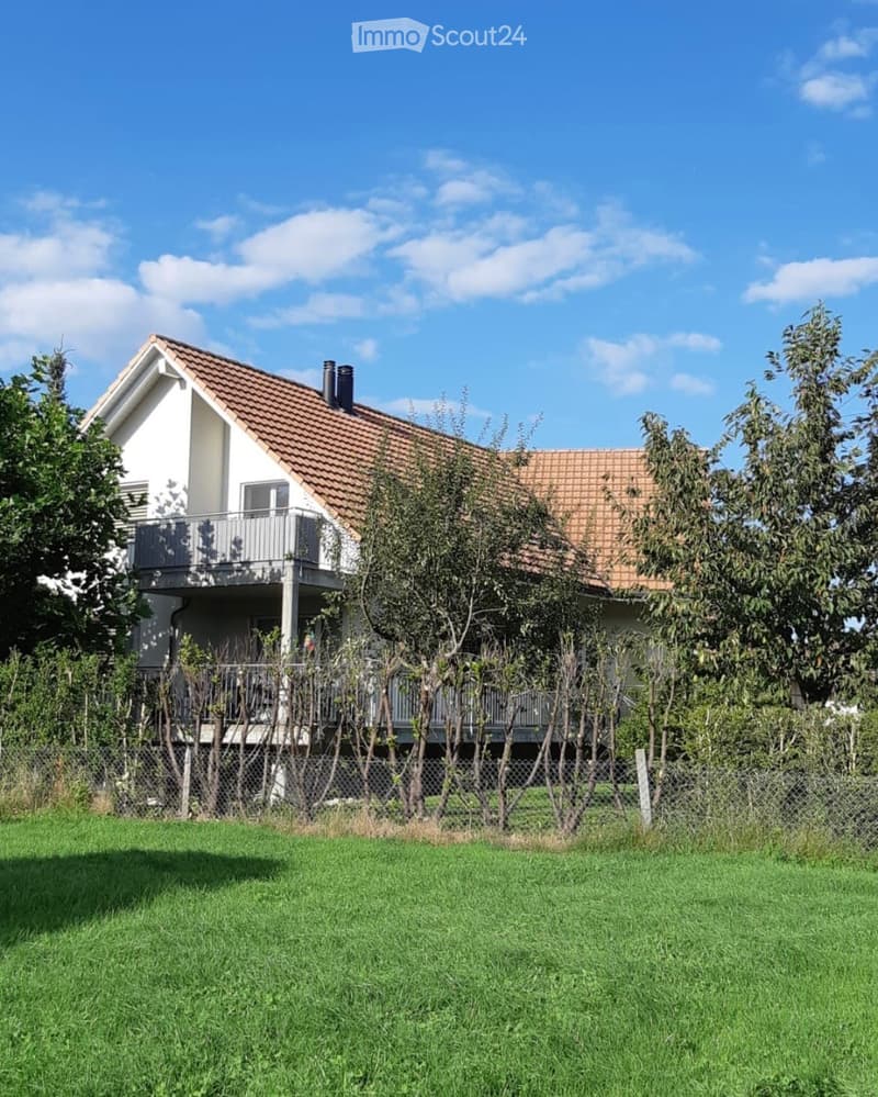 Two-story house with a tiled roof, white exterior, and a balcony on the upper level, surrounded by trees and a fenced yard.