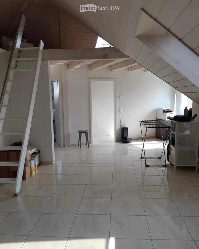 Bright, open room with slanted wooden ceiling and large windows. Features a small wooden stool, a metal table, a printer on a cabinet, and a staircase leading to another level.