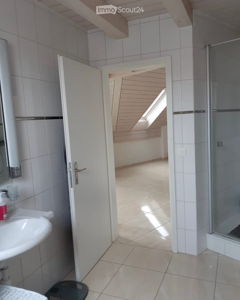Bathroom with white tiled walls, a sink, a mirror, a shower, and a candle on the sink counter. The door to the room is open, revealing a small, well-lit hallway with wooden flooring.