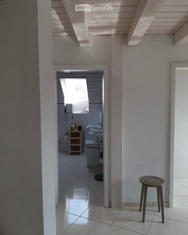 A modern hallway with white tiled floors, light-colored walls, and a small round stool placed near the wall. The hallway leads to a bathroom with a sink, mirror, and small wooden cabinet.