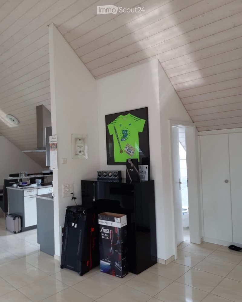 A modern, furnished room with a slanted wooden ceiling, white walls, and tiled floor. Visible objects include a black suitcase, a black speaker system, a black box, a framed jersey, a framed picture, a kitchen area with cabinets, a refrigerator, and vario