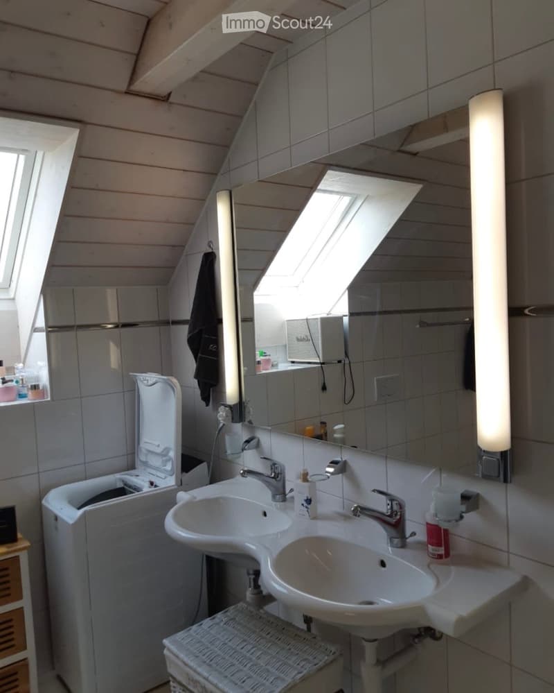 Modern bathroom with a double sink, large mirror, and built-in storage. Features include a skylight window, sleek fixtures, and a wicker stool.