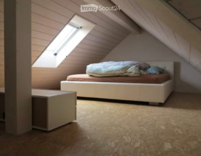 Cozy attic bedroom with a white bed, brown bedding, and a nightstand. The room features exposed wooden beams and a skylight window allowing natural light to fill the space.