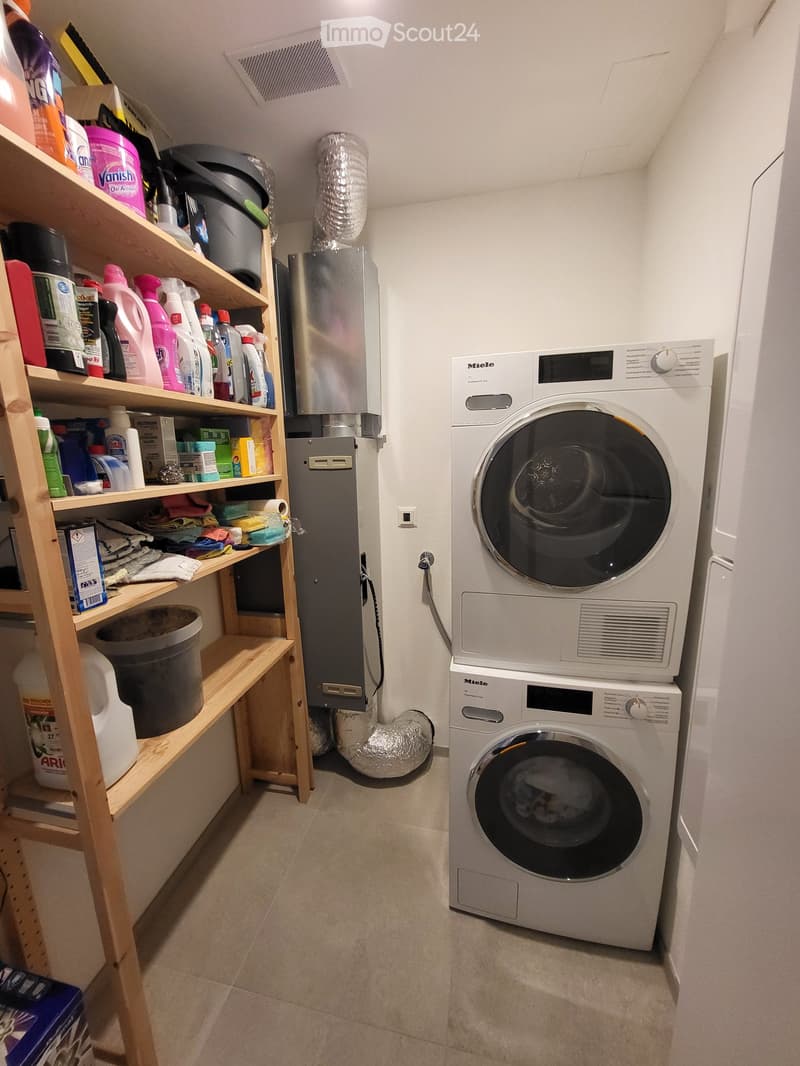 Laundry area with two stacked washing machines, wooden shelving unit with cleaning supplies, a plastic bin, a power outlet, and a ventilation duct.