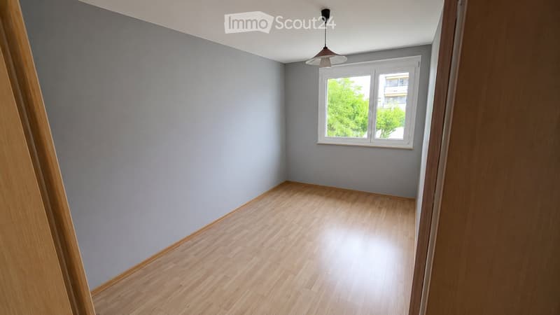 Empty room with light grey walls and wooden flooring, featuring a single window with white frame and a hanging light fixture.