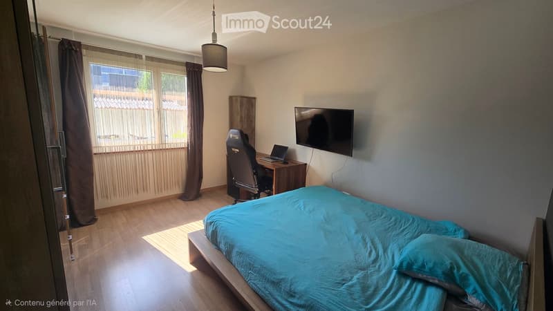 Modern bedroom with a wooden bed, a desk with a laptop, a chair, and a mounted screen on the wall. The room has wooden flooring, large windows with curtains, and a hanging light fixture.