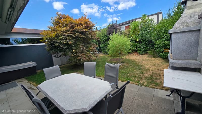 Outdoor patio area with a stone table, chairs, and a barbecue, surrounded by greenery and a clear blue sky.
