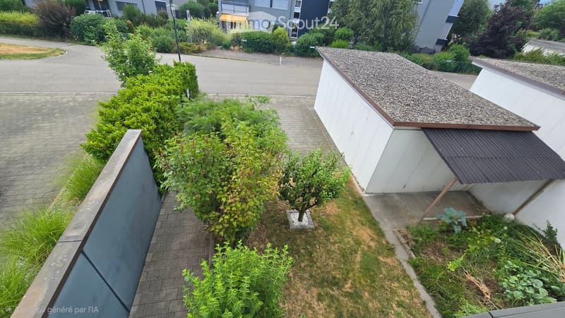 A small garden area with a tree, shrubs, and a paved pathway leading to a modern building with a gravel roof.
