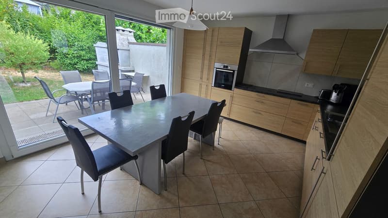 Modern kitchen and dining area with light wooden cabinets, stainless steel appliances, a large dining table with chairs, and a view to a patio with outdoor furniture.