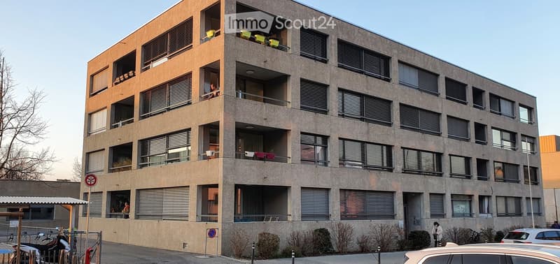 Modern multi-story apartment building with large windows, balconies, and visible furniture on some balconies.