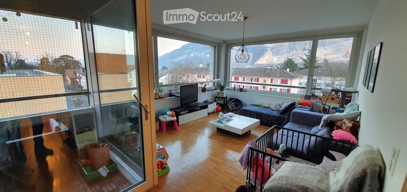 Bright living room with large windows offering a scenic mountain view. Features a sofa, coffee table, TV stand, a small pink stool, a cat tree, a baby crib, and various cat toys.